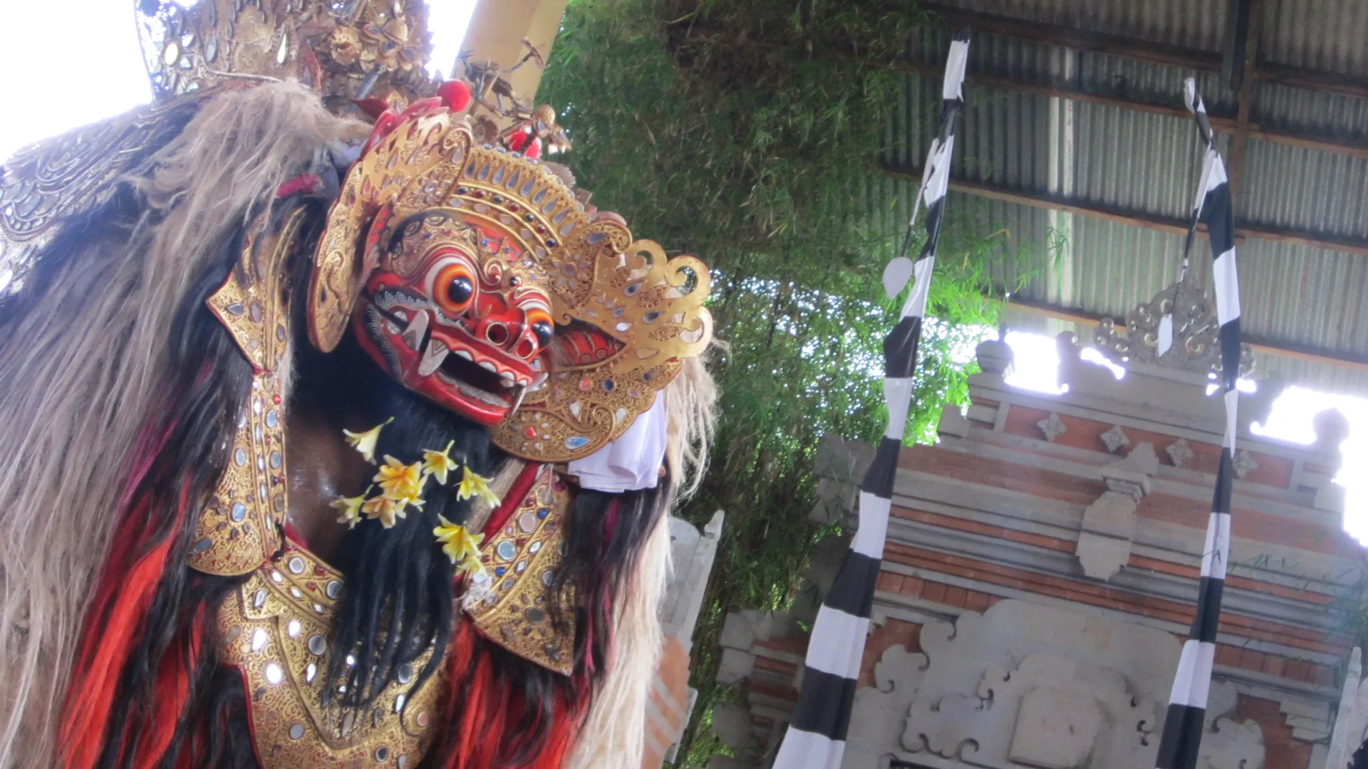Close-up of a Barong mask — the gold-crowned, red-faced lion-like guardian creature of Balinese dance — with a black-and-white poleng temple gate visible behind it
